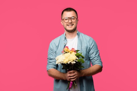 happy young man holding bouquet of flowers and smiling at camera isolated on pinkの写真素材