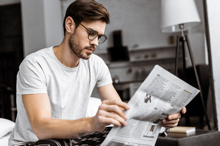 young man in pajamas and eyeglasses sitting on bed and reading newspaper in the morningの写真素材