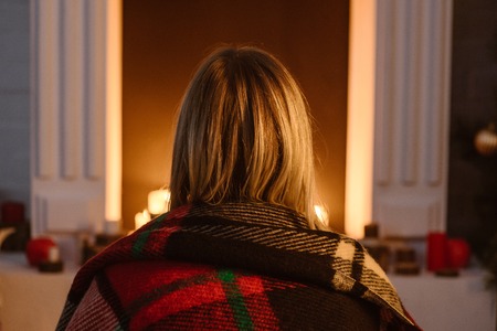 back view of woman in checkered blanket sitting near fireplace at home