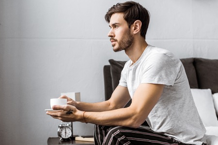 side view of young man in pajamas sitting on bed and holding cup of coffeeの写真素材