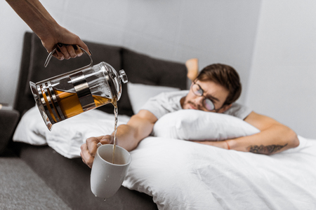 partial view of someone pouring tea into cup while sleeping man in eyeglasses lying on bedの写真素材