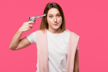 young woman holding revolver near head and looking at camera isolated on pink