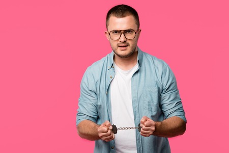aggressive young man in handcuffs and eyeglasses looking at camera isolated on pink