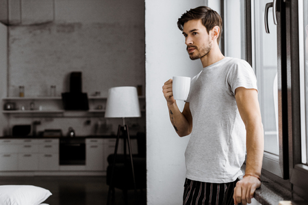 handsome young man in pajamas with mug of coffee leaning back on window at homeの写真素材