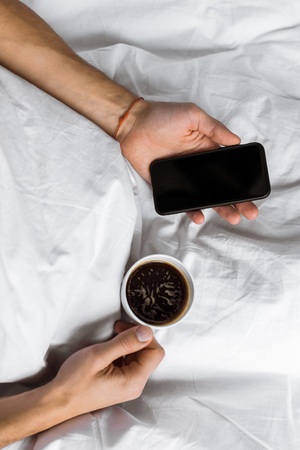 partial top view of man holding cup of coffee and smartphone with blank screen in bedの写真素材