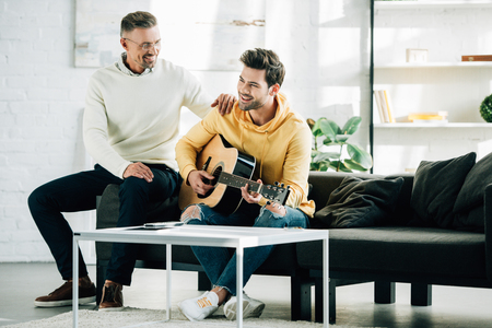 happy son playing acoustic guitar for mature father on weekend at homeの写真素材