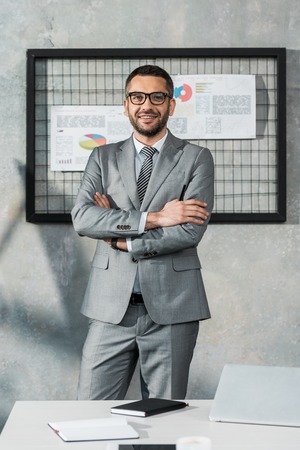 handsome businessman in suit and eyeglasses standing with crossed arms and smiling at camera in officeの写真素材
