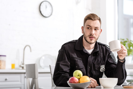 handsome policeman at kitchen sitting, holding cup of hot coffee and having breakfast at kitchen...