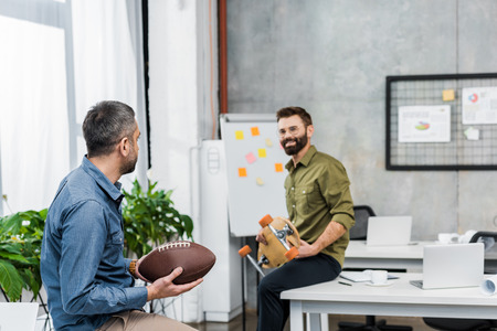 smiling businessmen looking at each other and holding american football ball and longboard in officeの写真素材