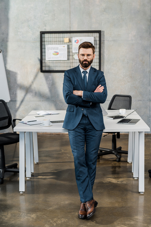 full length view of confident bearded businessman in suit and eyeglasses standing with crossed arms in officeの写真素材