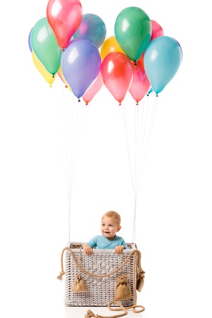 toddler boy smiling and standing in wicker basket with multicolored balloons isolated on whiteの写真素材