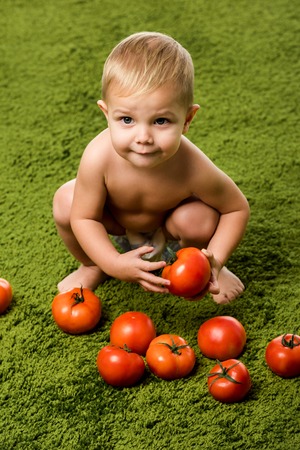 close up view of smiling toddler boy sitting on hunkers and holding tomato on green carpetの写真素材