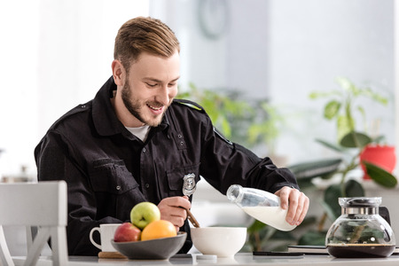 handsome policeman pouring milk into cereal bowl and having breakfast at kitchenの写真素材