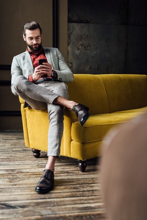 handsome stylish man using smartphone and sitting on yellow couchの写真素材
