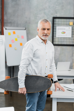 handsome middle aged businessman holding skateboard and looking at camera in officeの写真素材