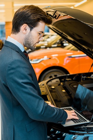 side view of businessman in eyeglasses checking car hood with laptop with blank screenの写真素材