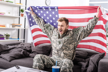 excited army soldier sitting on couch, cheering and proudly holding american flagの写真素材