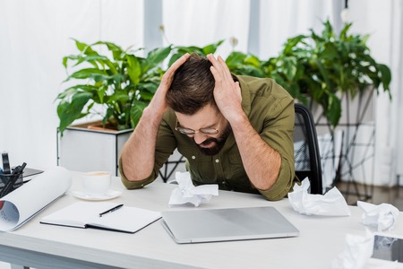tired handsome businessman touching head and sitting at table with crumpled paper in officeの写真素材
