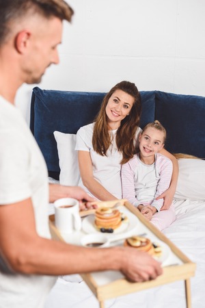 Father holding wooden tray with breakfast while daughter and wife sitting in bedの写真素材