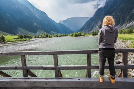 Back view of young woman standing on wooden bridge and looking at beautiful mountain river, Altai, Russiaの写真素材