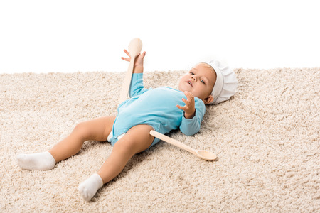 Toddler boy in chefs hat with two big wooden spoons lying on carpet and looking at camera isolated on whiteの写真素材