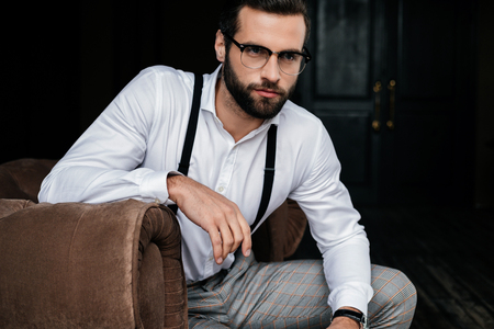 Handsome elegant man in eyeglasses, white shirt and suspenders sitting in armchairの写真素材