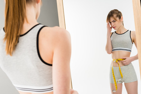 Young depressed woman measuring her waist and looking at mirror isolated on whiteの写真素材