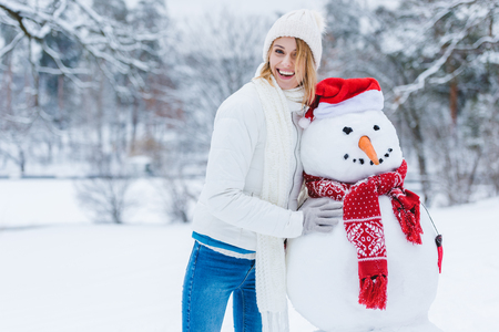 Portrait of cheerful young woman standing near snowman and looking at cameraの写真素材