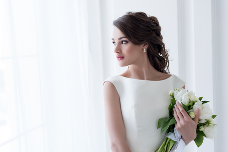 Beautiful bride holding wedding bouquet and looking at windowの写真素材