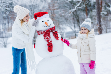 Happy mother and daughter making snowman together in winter forestの写真素材