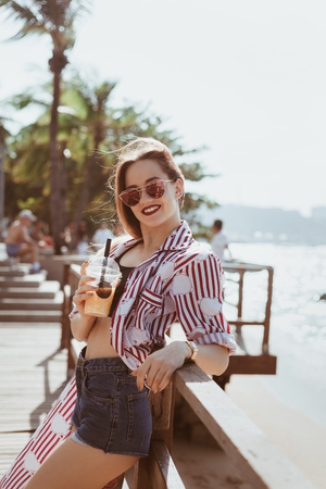 Smiling young woman with plastic cup of cocktail leaning back on pier at beachの写真素材