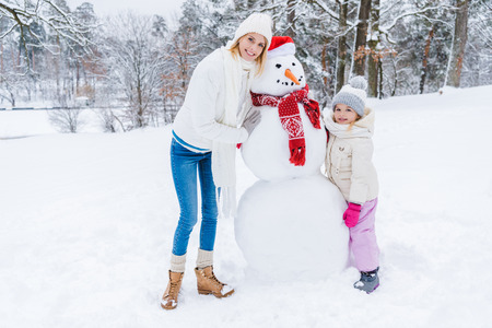 Beautiful happy mother and daughter smiling at camera while standing with snowman in winter parkの写真素材