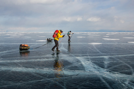 Two men with backpacks going through ice water surface and hills on background, Russia, Lake Baikalの写真素材