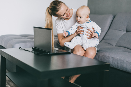 Beautiful smiling young mother holding adorable infant child while using laptop at homeの写真素材