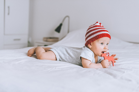 Infant child in striped red and white hat biting wooden deer decoration in bedの写真素材