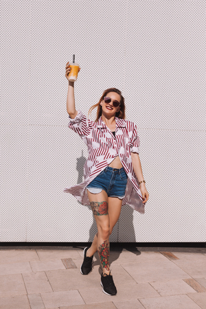 Smiling woman holding plastic cup of cocktail in front of white wallの写真素材