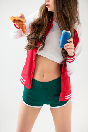 cropped image of sexy girl in baseball jacket holding pizza and can isolated on whiteの写真素材