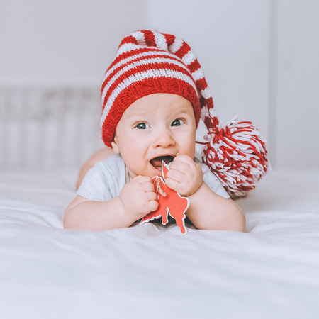 adorable infant child in red and white striped hat with pompom playing with toy angel in bedの写真素材