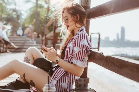 beautiful young woman using smartphone while sitting on pier at beachの写真素材