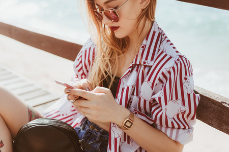 stylish young woman using smartphone outdoors while sitting on floor of pier in front of blurred seaの写真素材