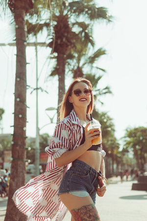 happy young woman drinking mango shake on sunny street with palm treesの写真素材