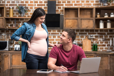 smiling young man using laptop and looking at pregnant wife at homeの写真素材