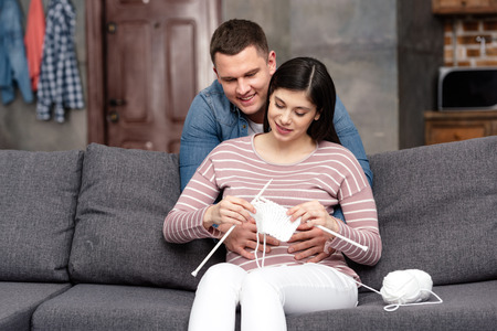 smiling young man hugging pregnant woman sitting on sofa and knitting at homeの写真素材