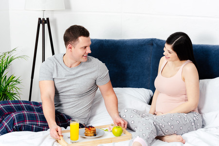 smiling young man holding tray with breakfast and looking at happy pregnant woman in bedの写真素材