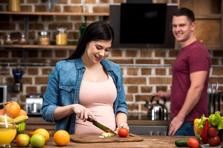 happy young pregnant woman cutting vegetables while smiling husband standing behind in kitchenの写真素材