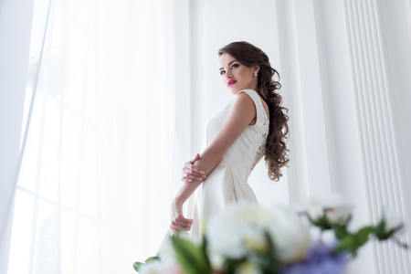 Brunette caucasian bride in wedding dress at window, flowers on foregroundの写真素材