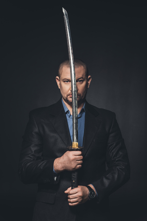 Mature man in suit holding Japanese katana sword in front of his face isolated on blackの素材 [FY310112770144]