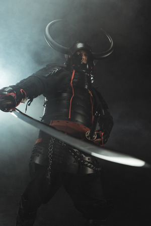 Bottom view of samurai in traditional armor with sword on dark background with smokeの写真素材