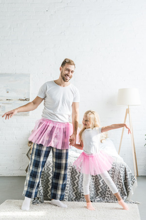 happy father and adorable little daughter in pink tutu skirts holding hands and dancing at homeの写真素材