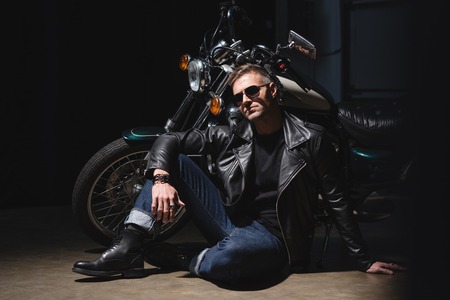 handsome biker in sunglasses standing by motorcycle in garage